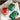 Three Christmas-themed bath products arranged on a white towel: a red ornament-shaped pump bottle, a green ornament-shaped pump bottle, and a green rectangular package with a bow. The products feature holiday patterns and gold pump hardware.